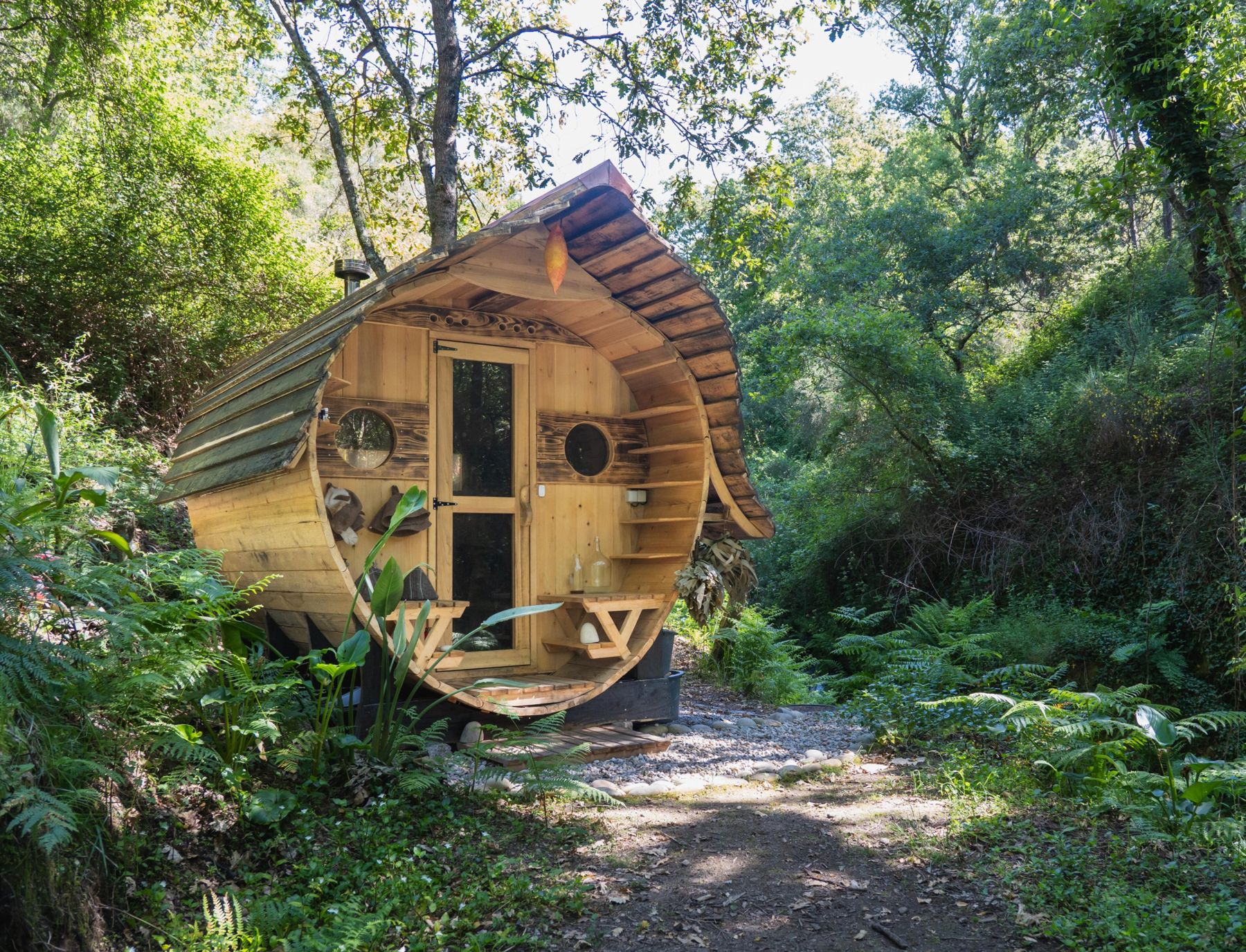The barrel sauna in the forest by the creek, with porthole windows and a curved shingle roof